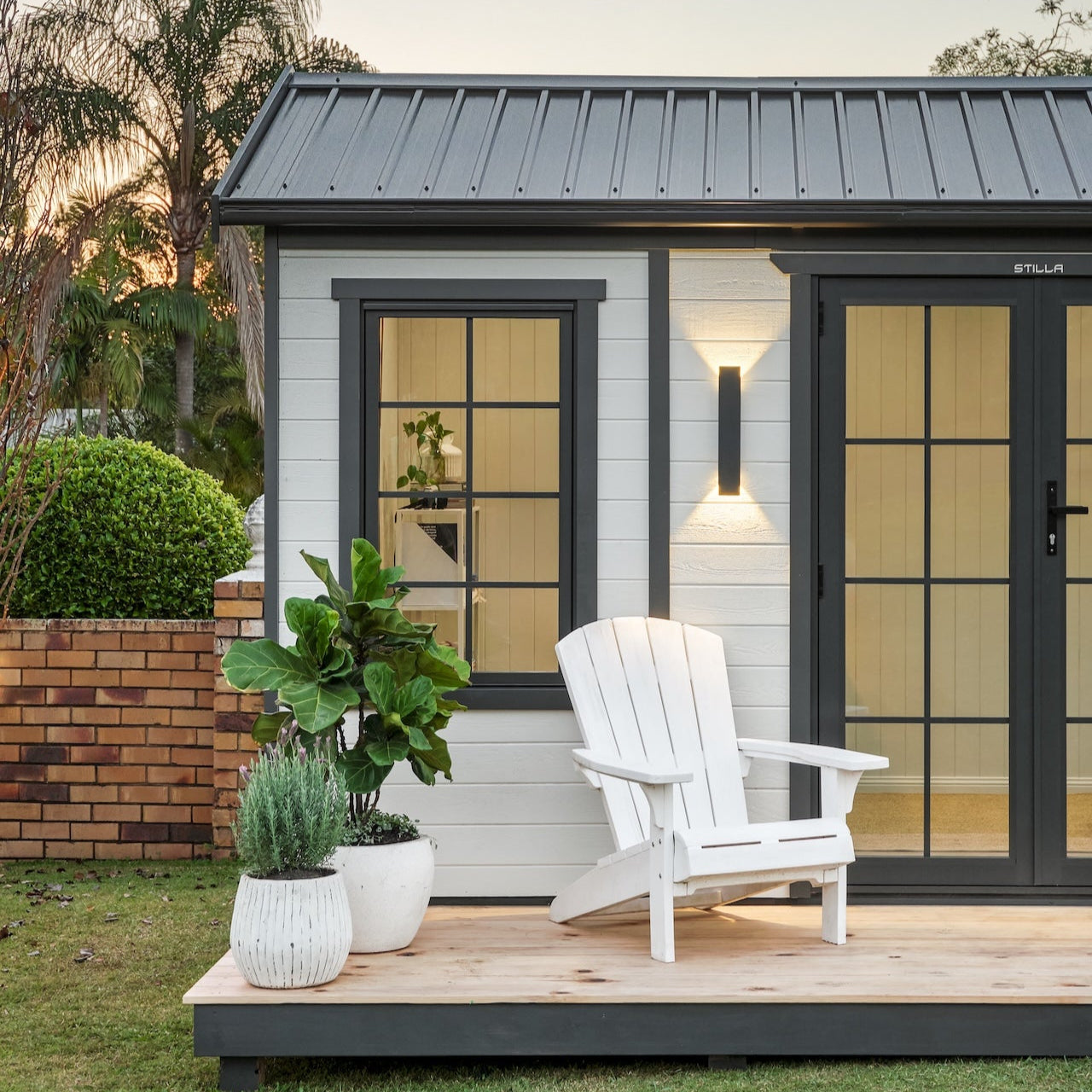 Small house with a white chair and plants on a wooden deck at sunset.