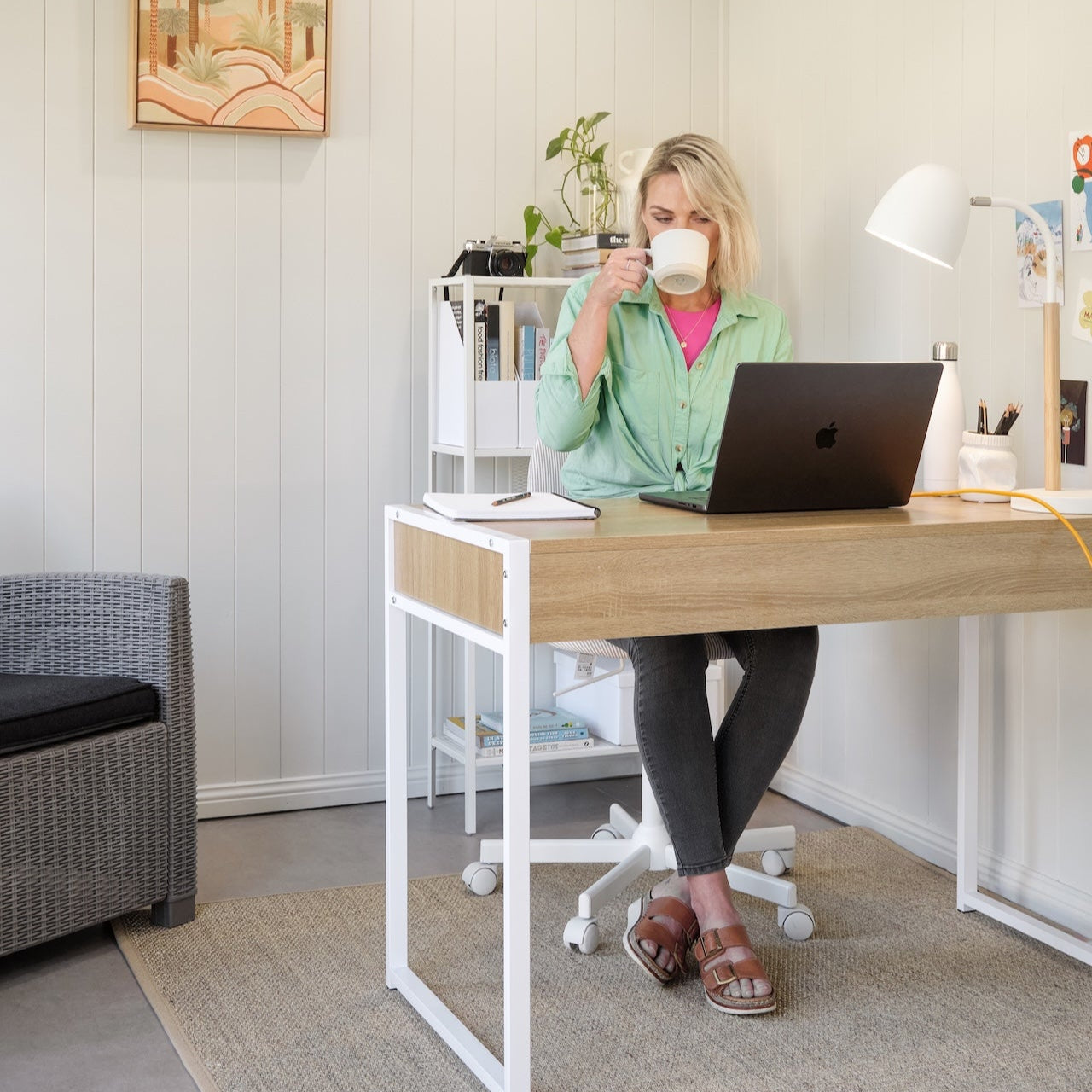Person sitting at a desk with a laptop, drinking from a mug in a home office setting.