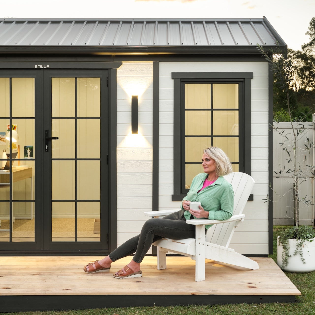 Woman sitting on a chair in front of a modern shed with a sunset sky.